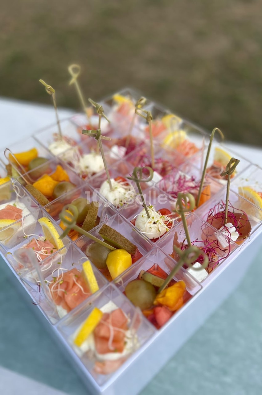 Plateau de mises en bouche individuelles avec fruits, charcuterie et décoration florale sur piques dorées.