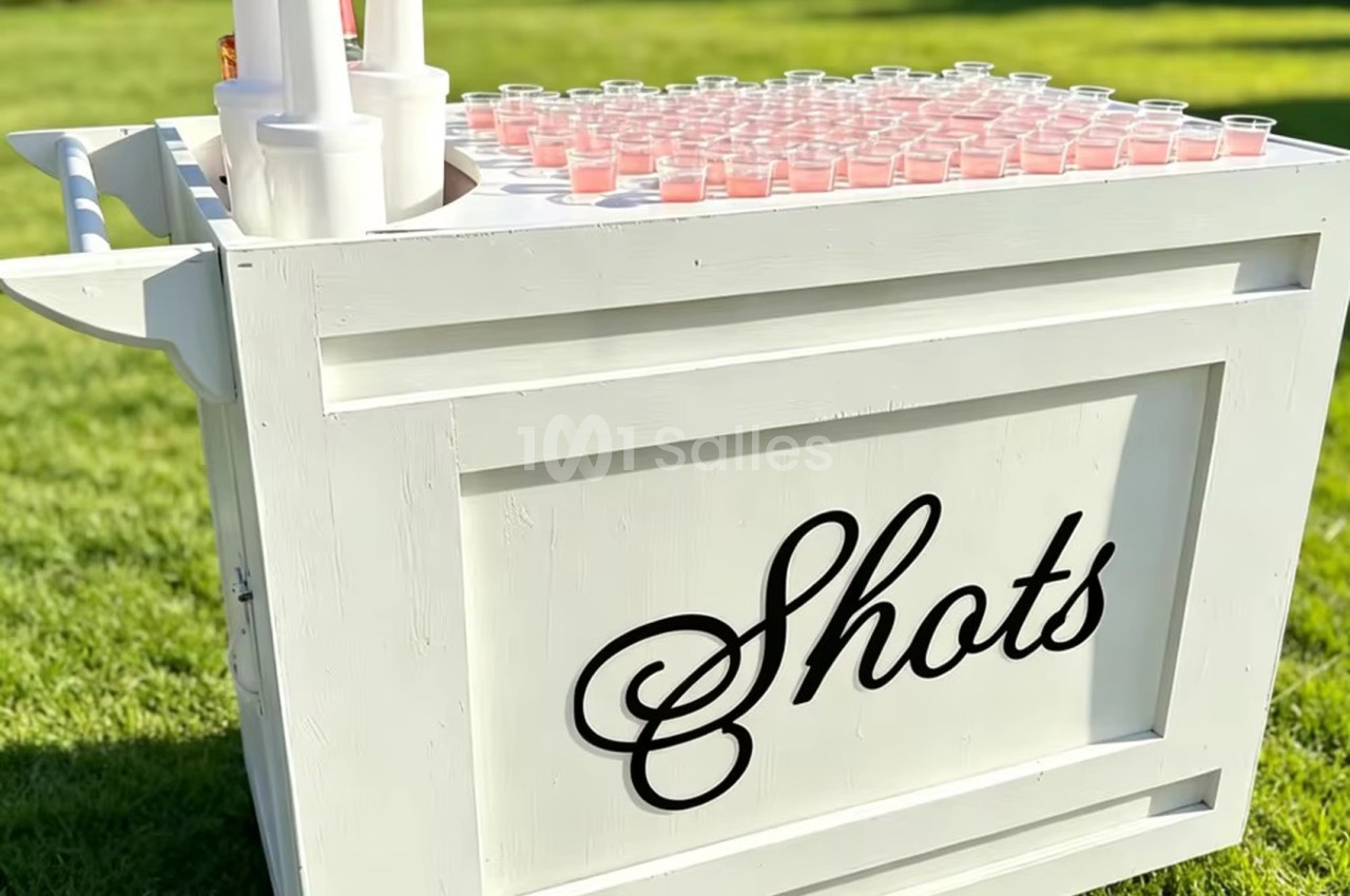 Comptoir blanc en bois avec verres de shots rose pâle alignés dessus, dans un jardin.