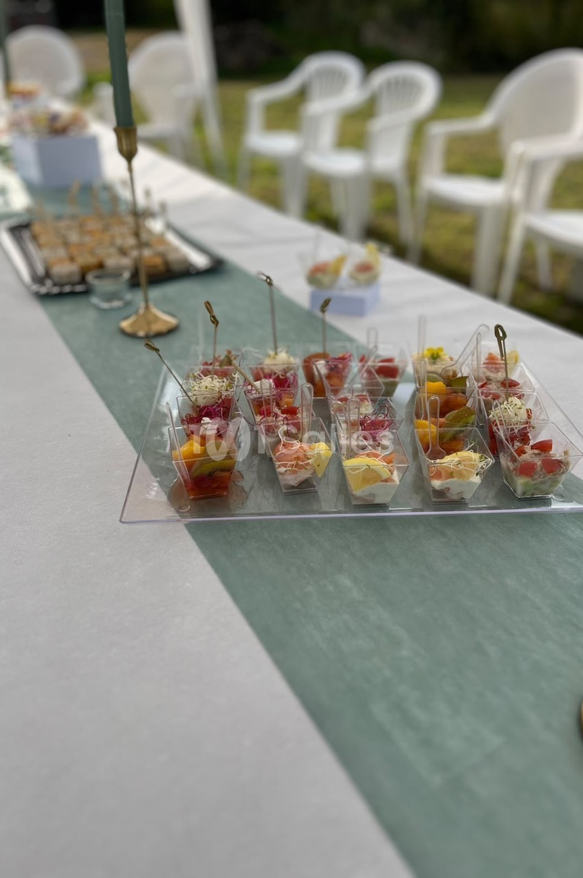 Table dressée en extérieur avec amuse-bouches en verrines garnies de charcuterie et fruits - chaises blanches visibles en arrière-plan.