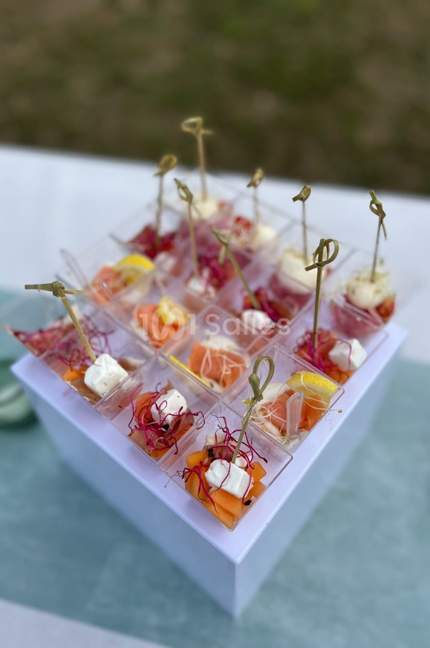 Plateau blanc présentant des canapés individuels garnis de fromage frais, saumon et légumes, piqués de tiges dorées.