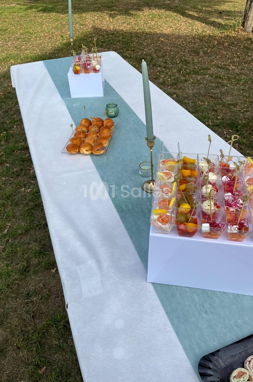 Table dressée en extérieur avec nappe blanche et bleu ciel, petit-fours, verre, bougies et verres de cocktails décorés aux fruits.