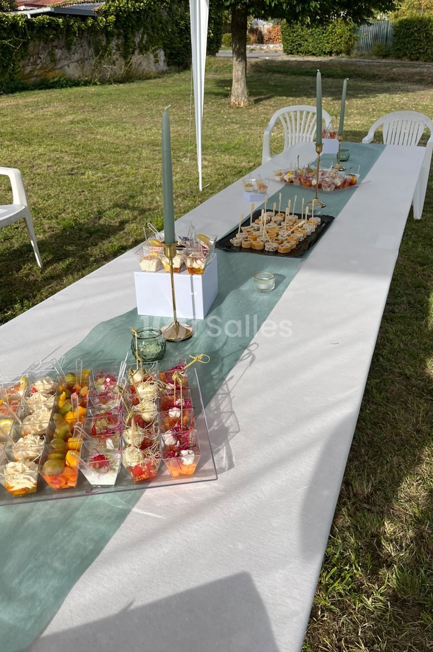 Table dressée en extérieur avec nappe blanche et chemin vert, canapés variés, coupes de fruits frais et candélabres dorés.