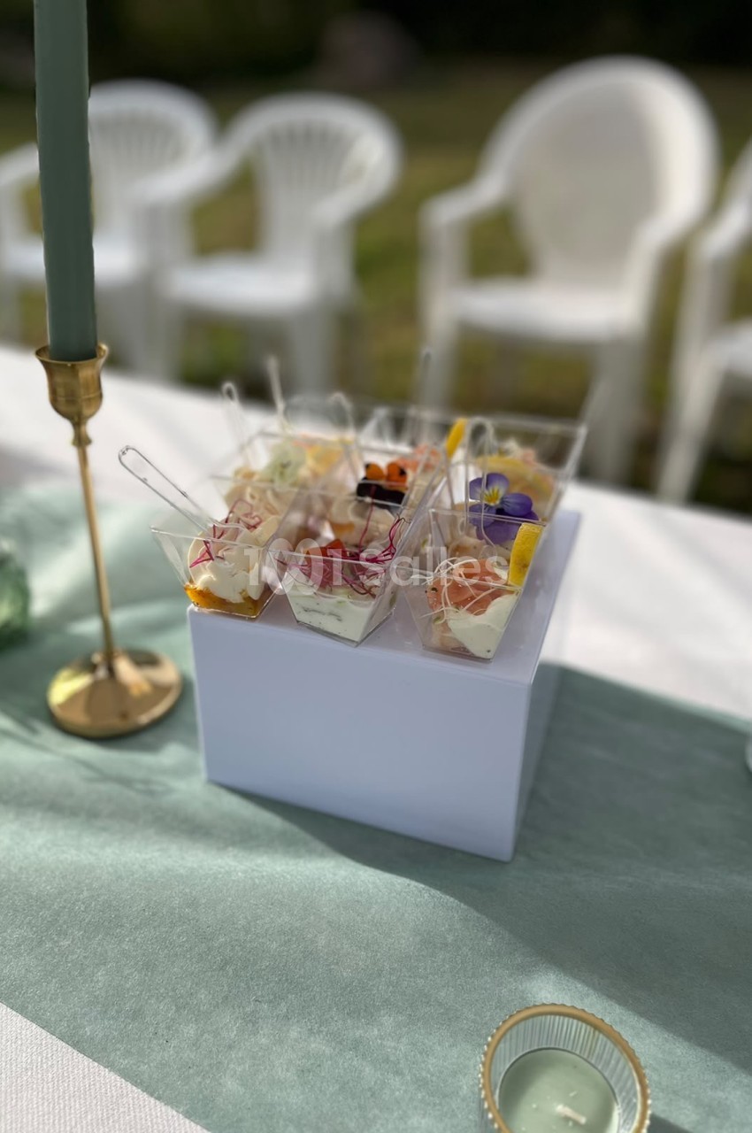 Présentoir blanc de petits fours individuels en verrine avec garnitures colorées, bougeoir doré et chaises blanches en arrière-plan.