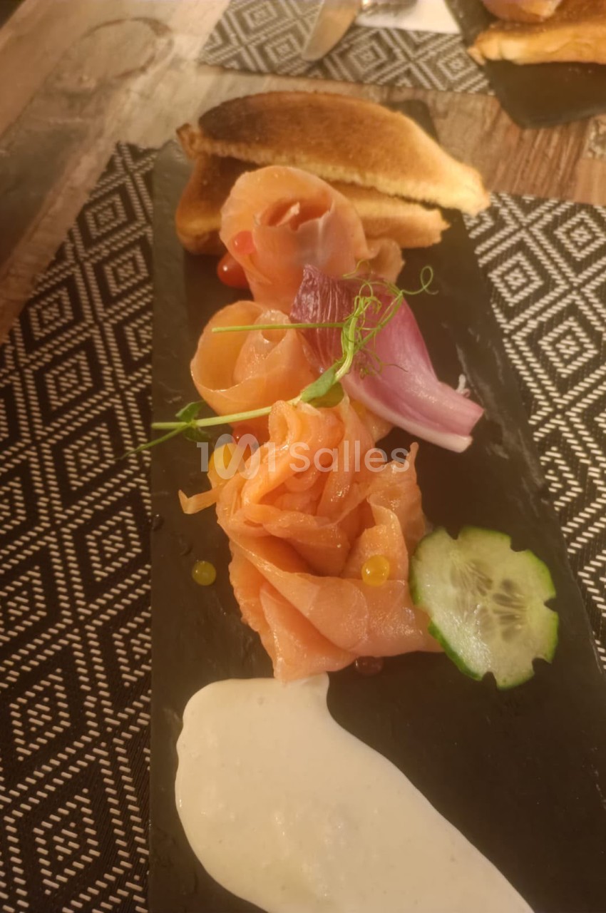 Assiette de saumon fumé roulé, fleur comestible rose et tranche de concombre sur socle blanc.