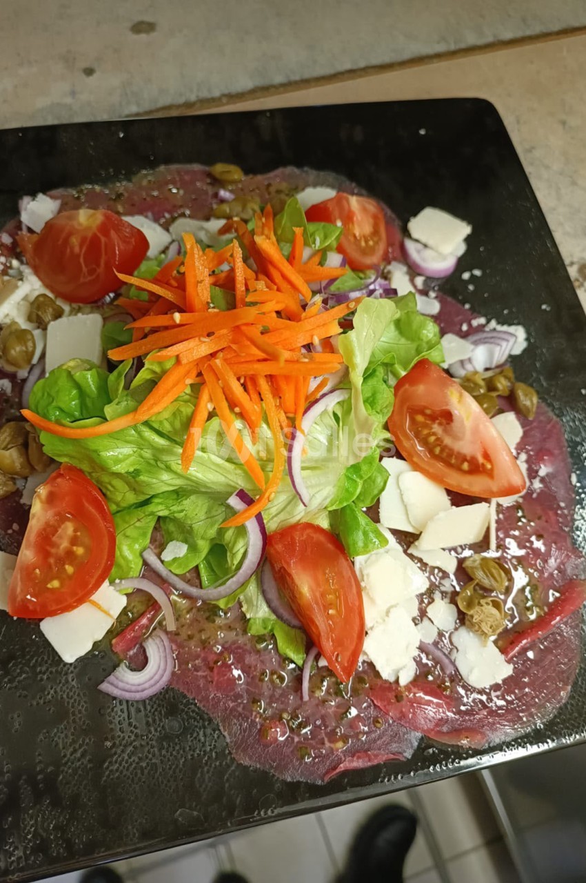 Assiette noire présentant une carpaccio de viande rouge garnie de roquette, tomates, carottes râpées, fromage blanc et cornichons.