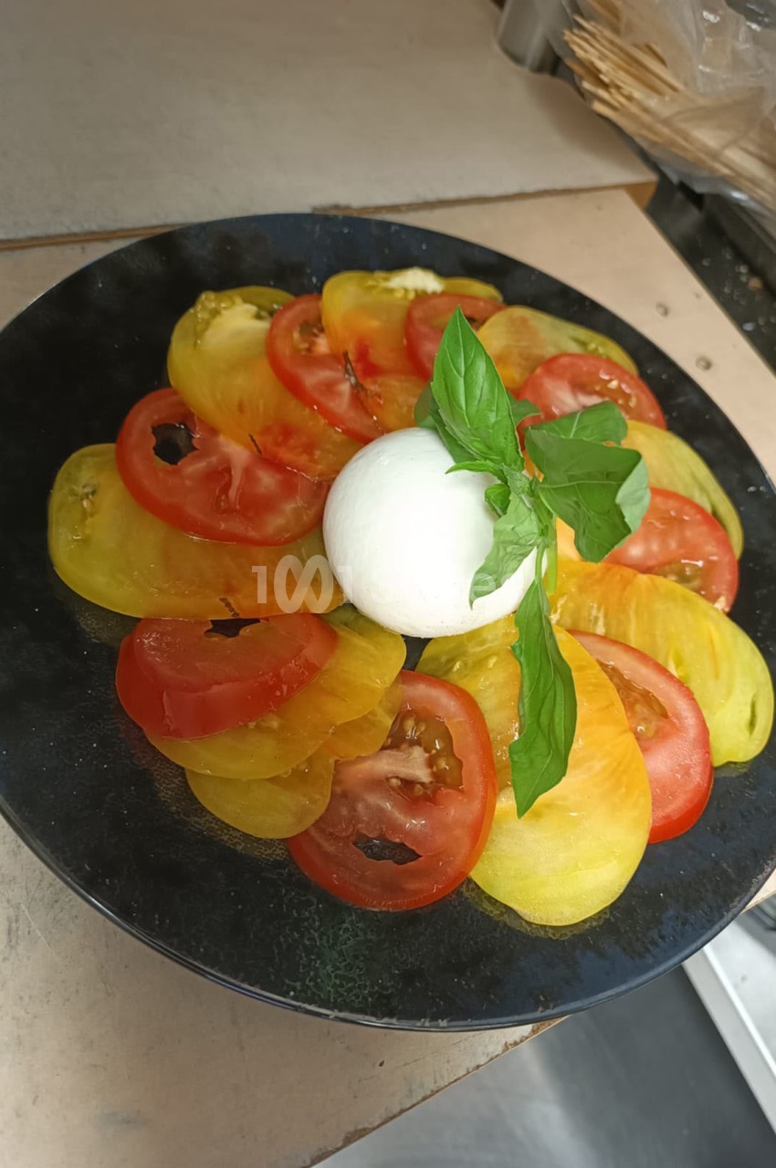 Salade caprese composée de tranches de tomates rouges et jaunes alternées, mozzarella fraîche et feuilles de basilic sur assiette noire.