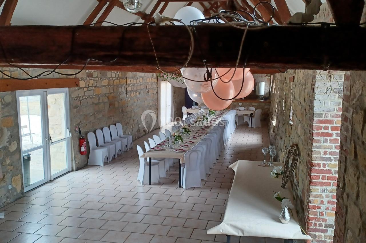 Salle en pierre avec une longue table décorée pour un événement, chaises blanches et ballons suspendus au plafond.