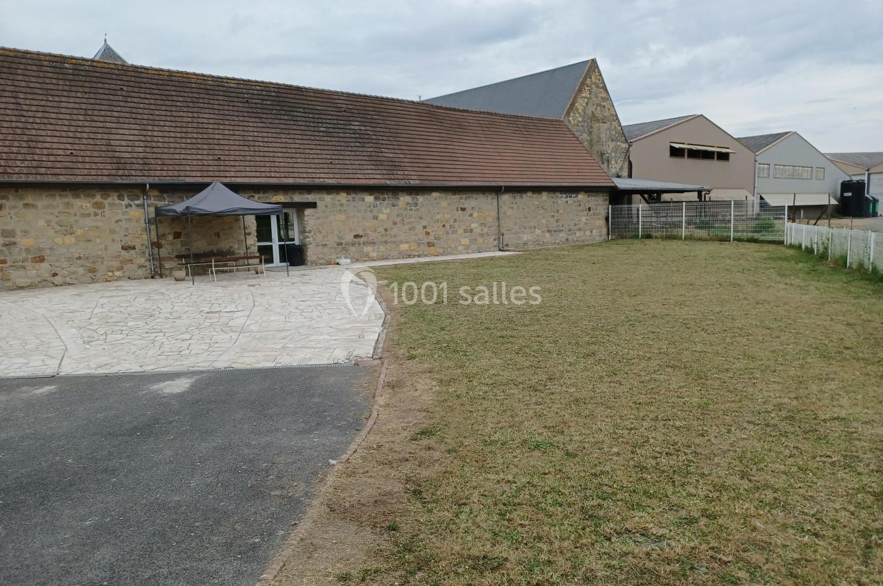 Bâtiment en pierre avec toit en tuiles, espace pavé à l'avant et pelouse sur le côté, ciel nuageux.