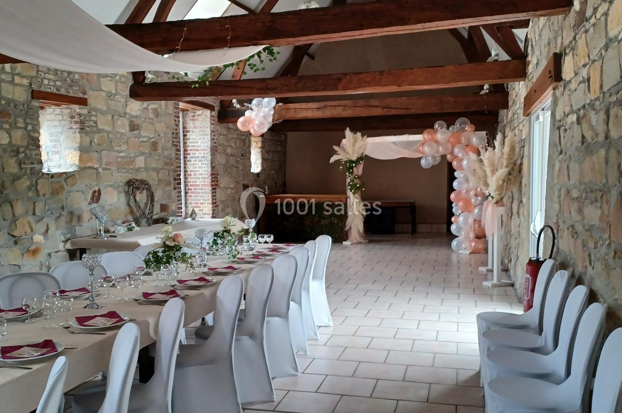 Salle de réception en pierre avec tables dressées, chaises blanches et décoration florale et ballons.