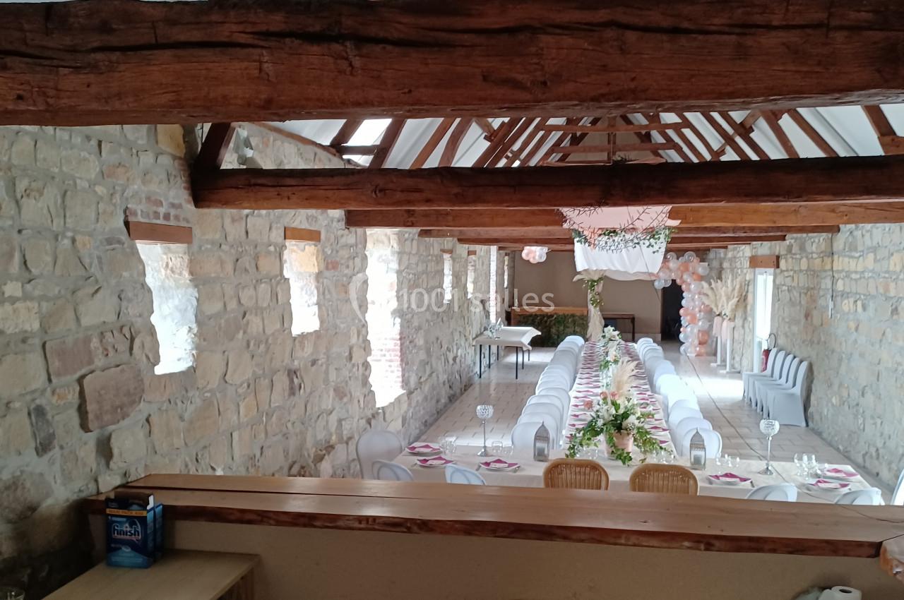 Salle en pierre avec poutres apparentes, décorée pour un événement, tables alignées avec nappes blanches et ornements…