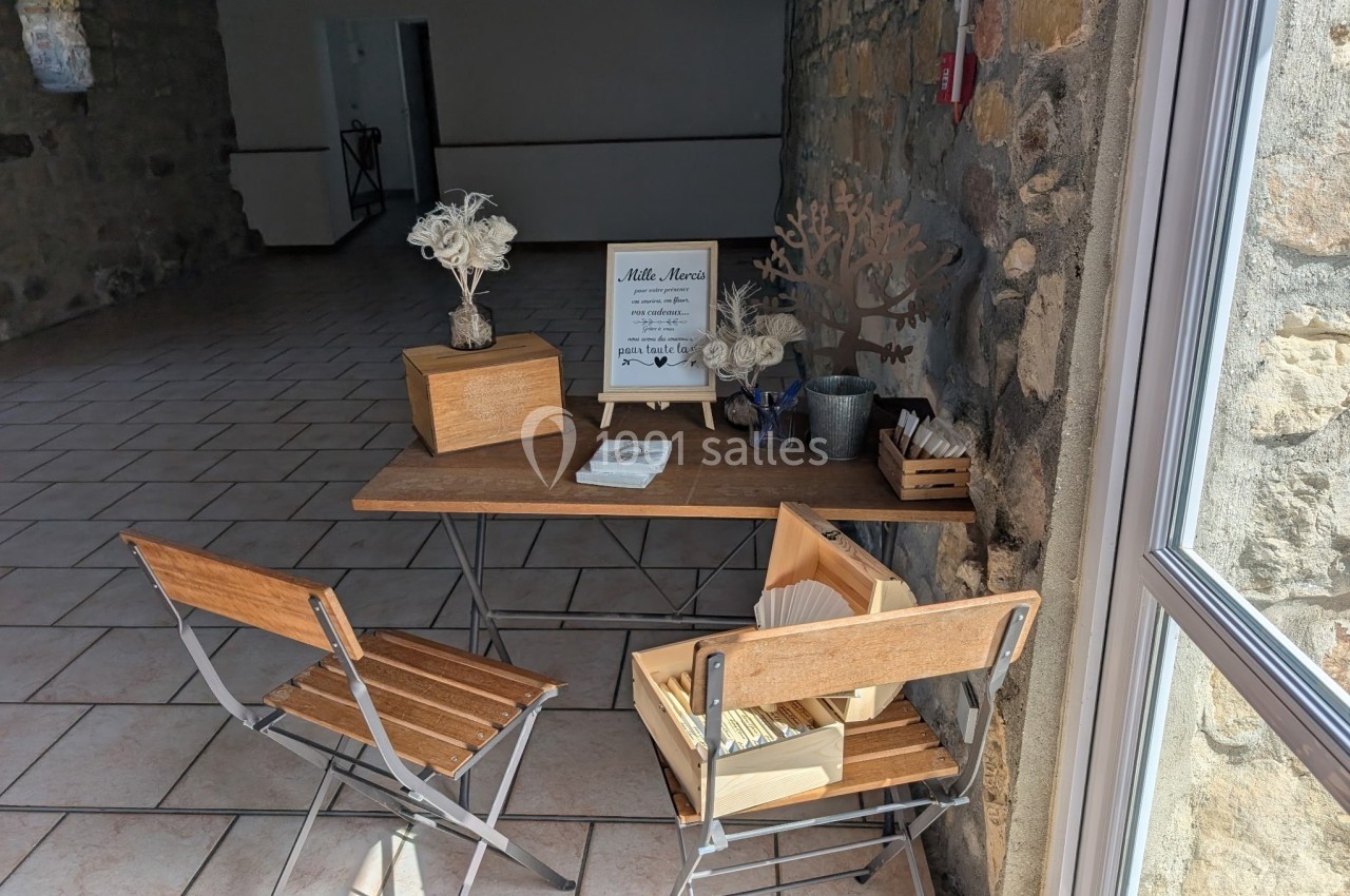 Table en bois avec deux chaises, décorée de fleurs séchées et d'un panneau ’Livre d'or’, dans une salle lumineuse en pierre.