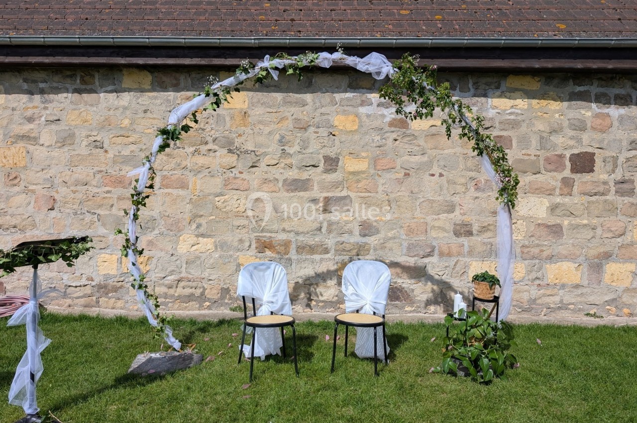 Arche décorée de feuillage et de tissu blanc avec deux chaises recouvertes, installées sur une pelouse devant un mur en…