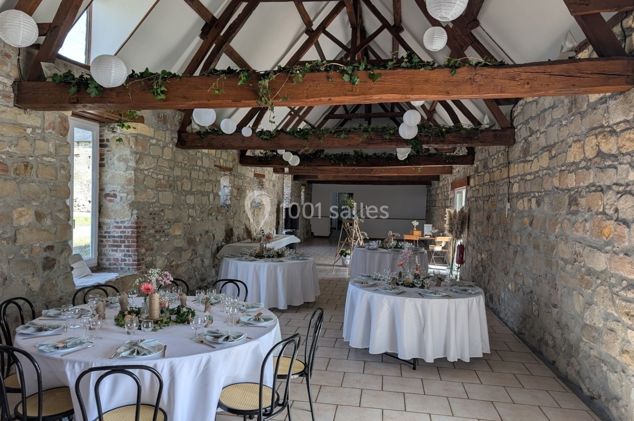 Salle en pierre avec poutres apparentes, tables rondes dressées pour un événement, décorée de guirlandes et lanternes…