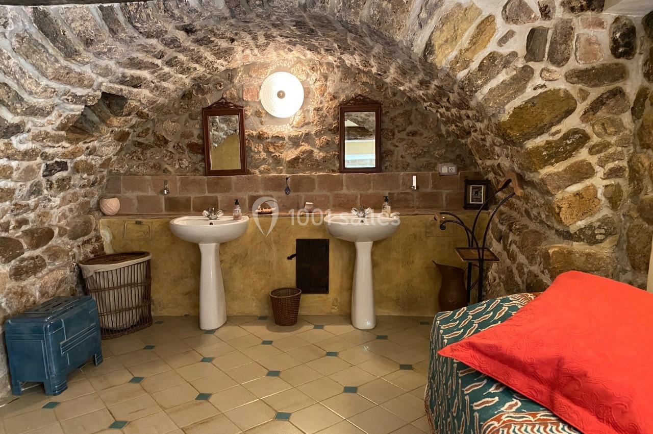 Salle de bain rustique avec murs en pierre voûtés, deux lavabos, un lit avec couvre-lit rouge et un sol carrelé.