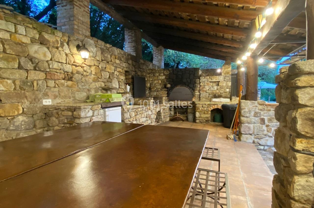 Cuisine extérieure en pierre avec un four à bois, un plan de travail, un éclairage suspendu et une grande table en métal.