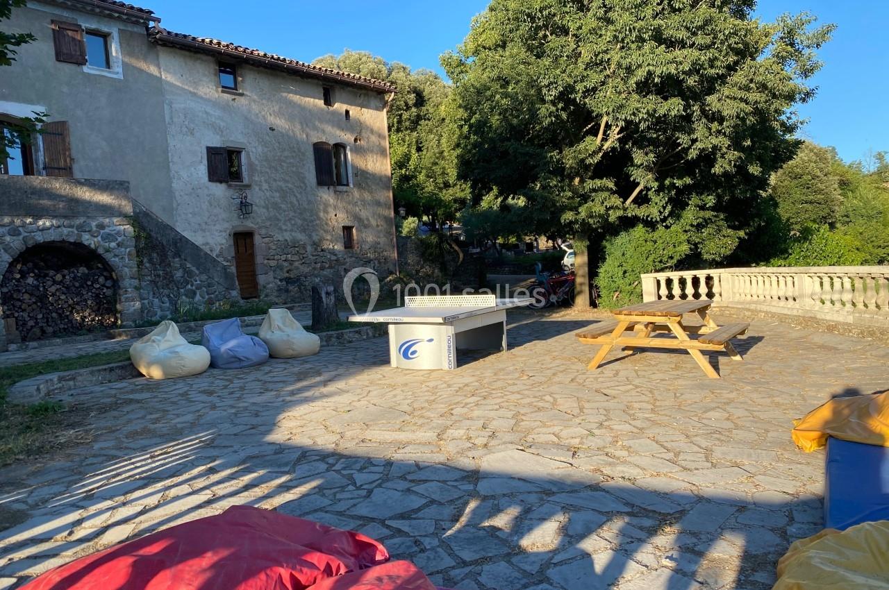 Cour ensoleillée avec table de ping-pong, bancs en bois, poufs colorés et maison en pierre entourée de verdure.