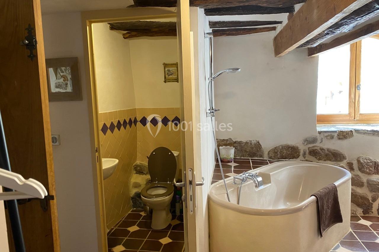 Salle de bain rustique avec baignoire d'angle, toilettes séparées, murs en pierre et poutres apparentes.