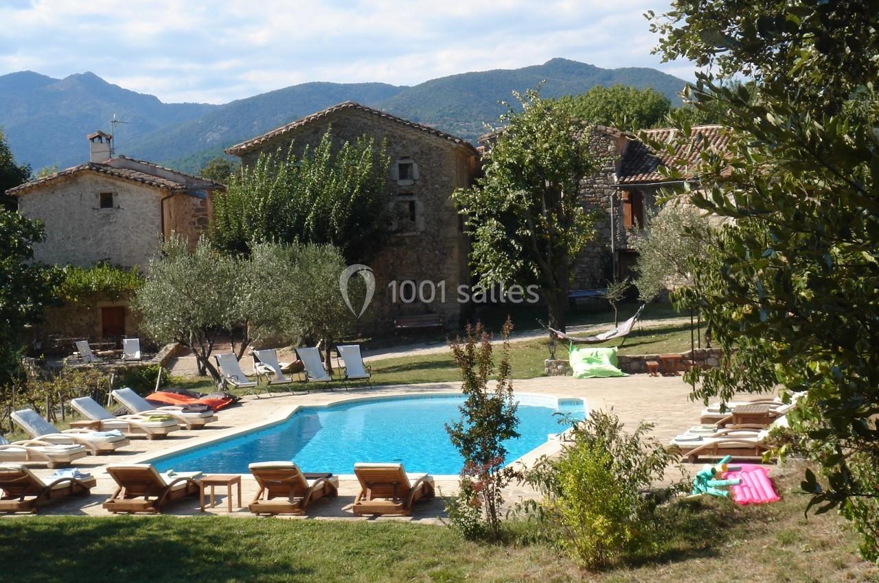 Piscine extérieure entourée de chaises longues, arbres et maisons en pierre, avec des montagnes en arrière-plan.