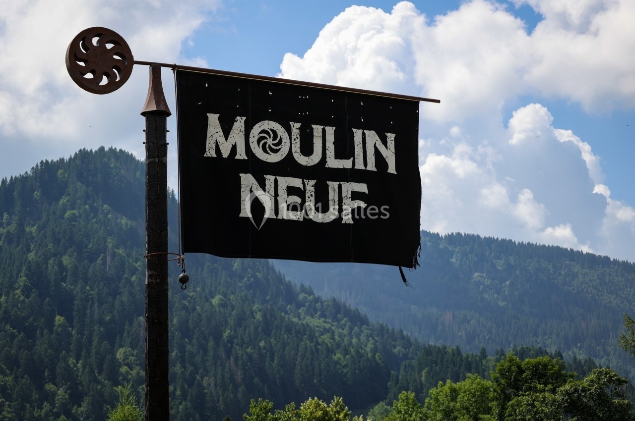 Bannière noire avec l'inscription ’Moulin Neuf’ suspendue à un poteau, devant un paysage de montagnes et de forêt.