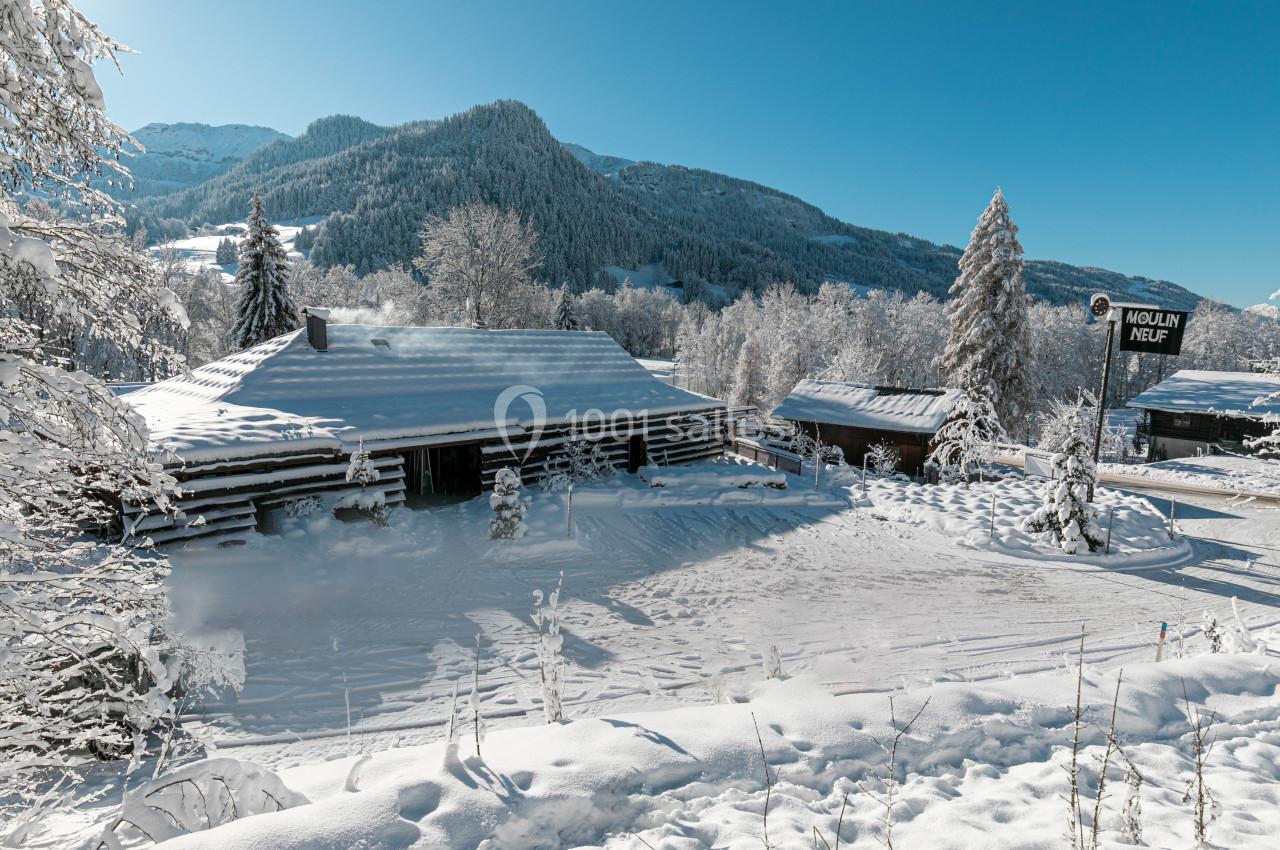 Chalet en bois entouré de neige, avec des montagnes et des arbres enneigés en arrière-plan sous un ciel bleu.