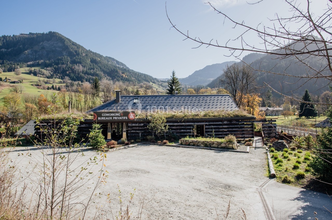 Bâtiment en bois entouré de montagnes et de végétation, avec un parking gravillonné à l'avant.