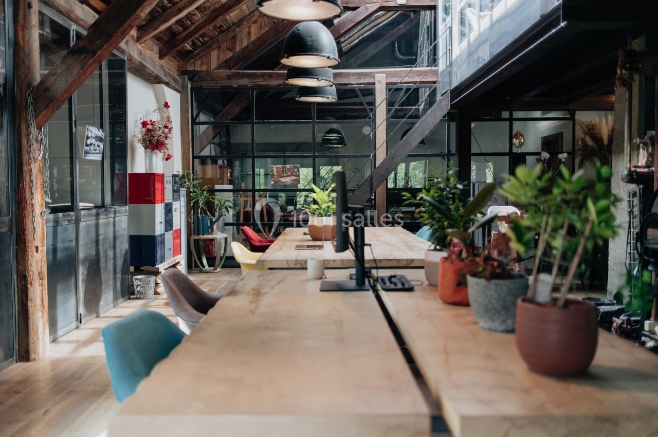 Espace de travail lumineux avec de longues tables en bois, des plantes en pot et un plafond en poutres apparentes.