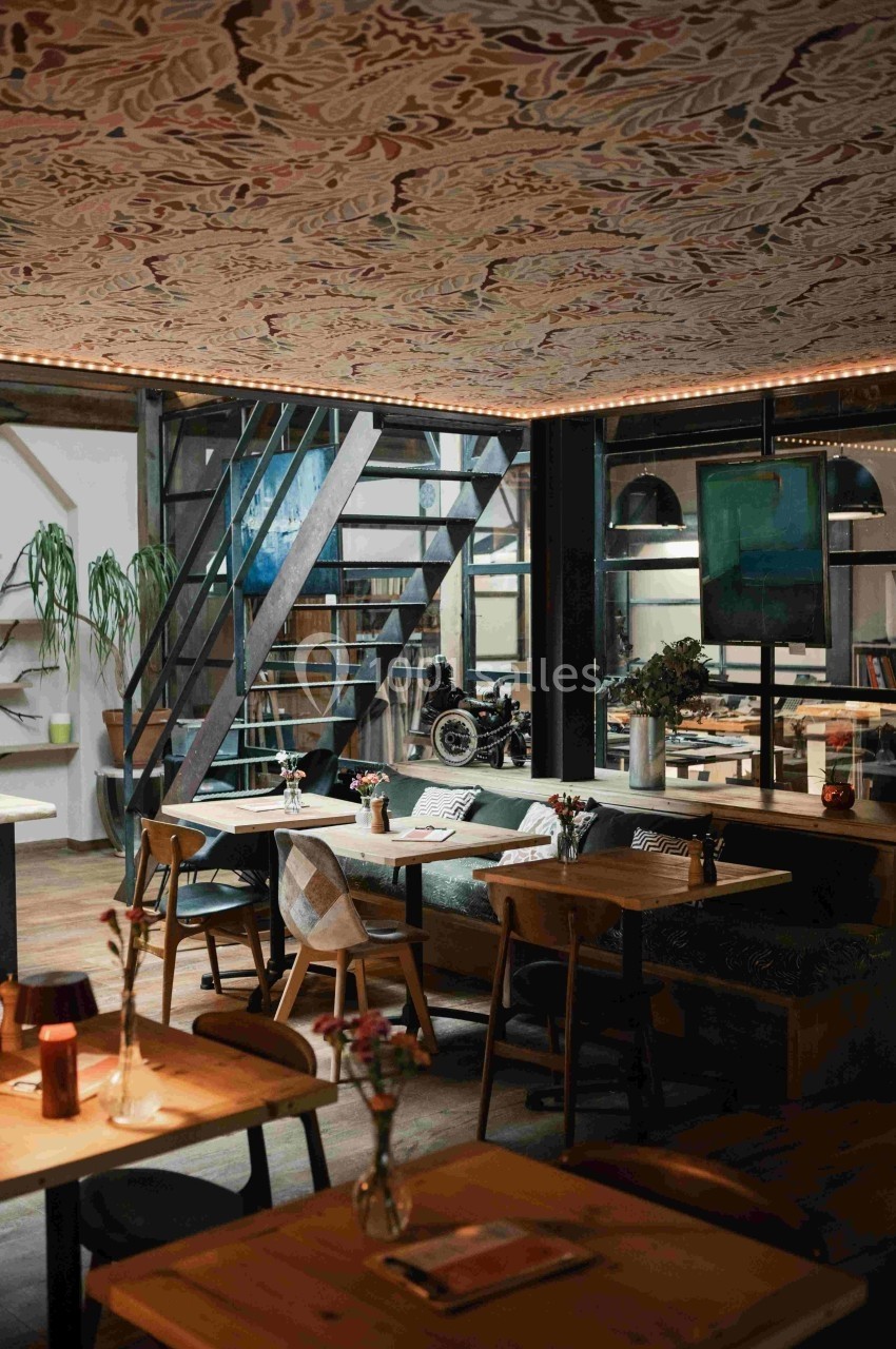 Salle de restaurant moderne avec tables en bois, banquettes, escalier métallique et plafond décoré.