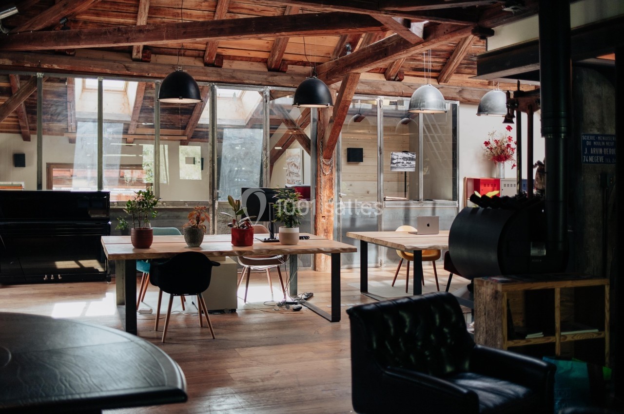 Espace de travail lumineux avec poutres apparentes, tables en bois, plantes et chaises dans un style industriel.