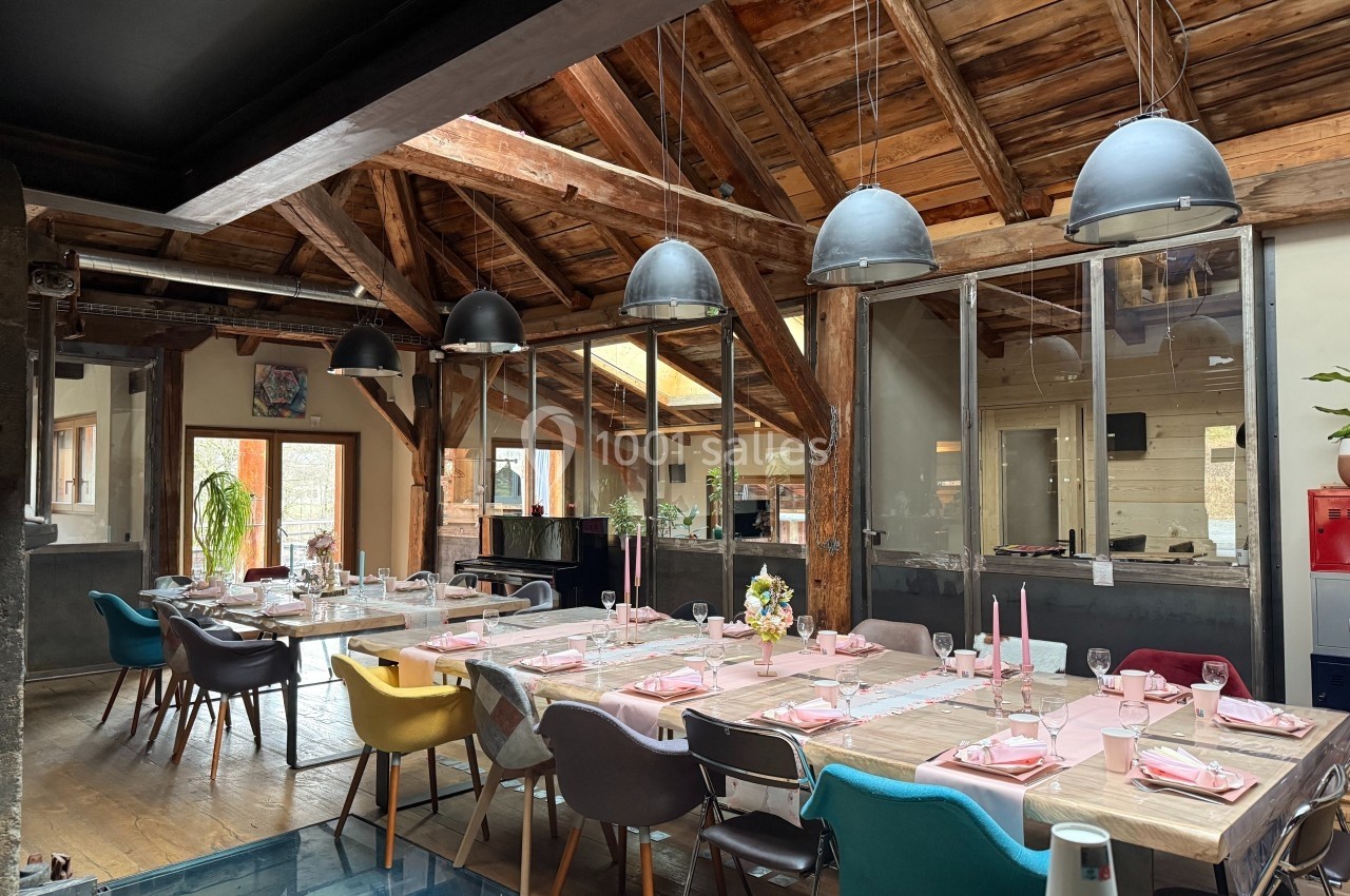 Salle à manger lumineuse avec grande table dressée, chaises colorées et plafond en bois avec poutres apparentes.