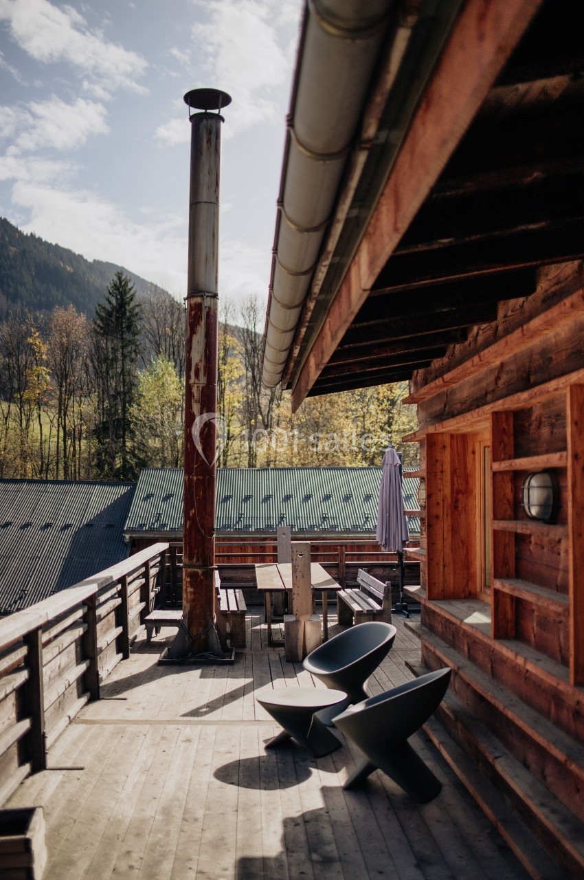 Terrasse en bois d'un chalet avec mobilier extérieur, cheminée métallique et vue sur des montagnes et arbres.