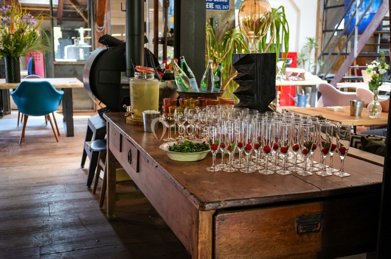 Table en bois avec verres à pied, carafe de boisson, herbes fraîches et décor intérieur lumineux et végétalisé.