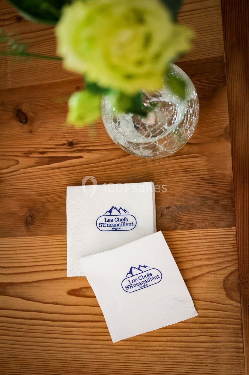 Deux serviettes blanches avec un logo bleu posées sur une table en bois, près d'un vase contenant des fleurs vertes.
