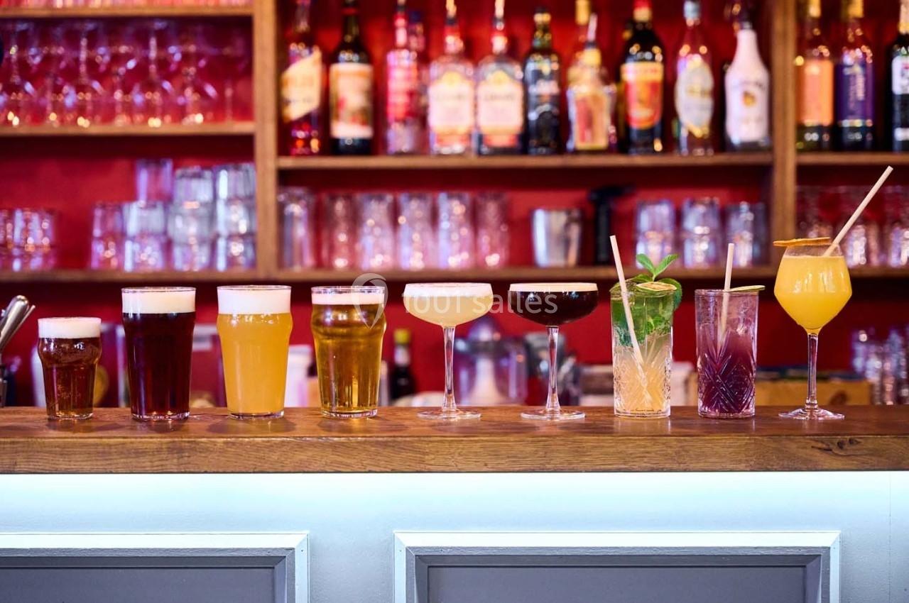 Alignement de verres contenant diverses boissons colorées sur un comptoir de bar, avec des bouteilles en arrière-plan.