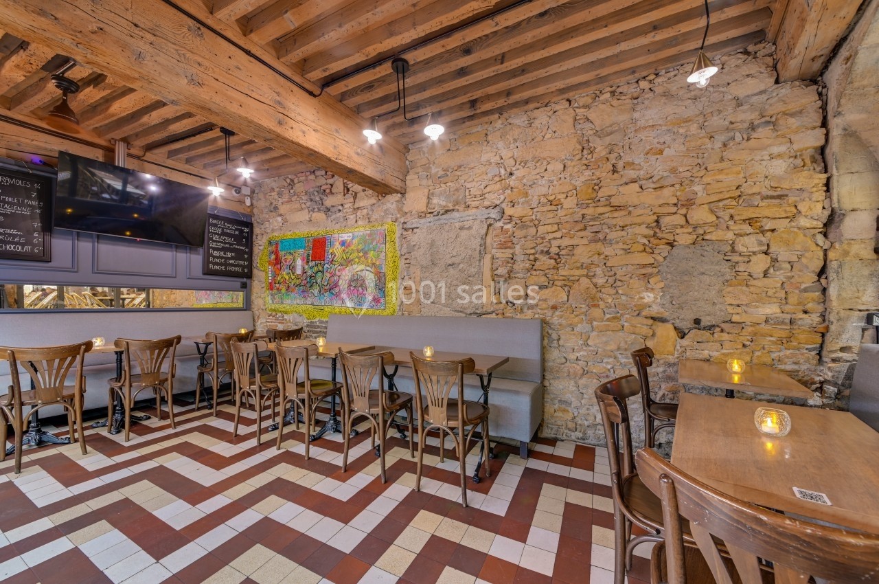 Salle de restaurant avec murs en pierre, plafond en bois, tables en bois et banquettes, éclairée par des lampes suspendues.