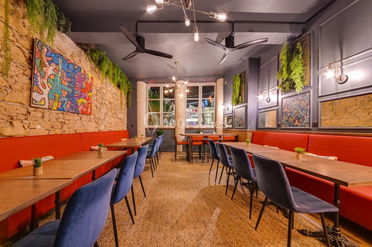 Salle de restaurant lumineuse avec murs en pierre, banquettes rouges, chaises bleues et décoration végétale.