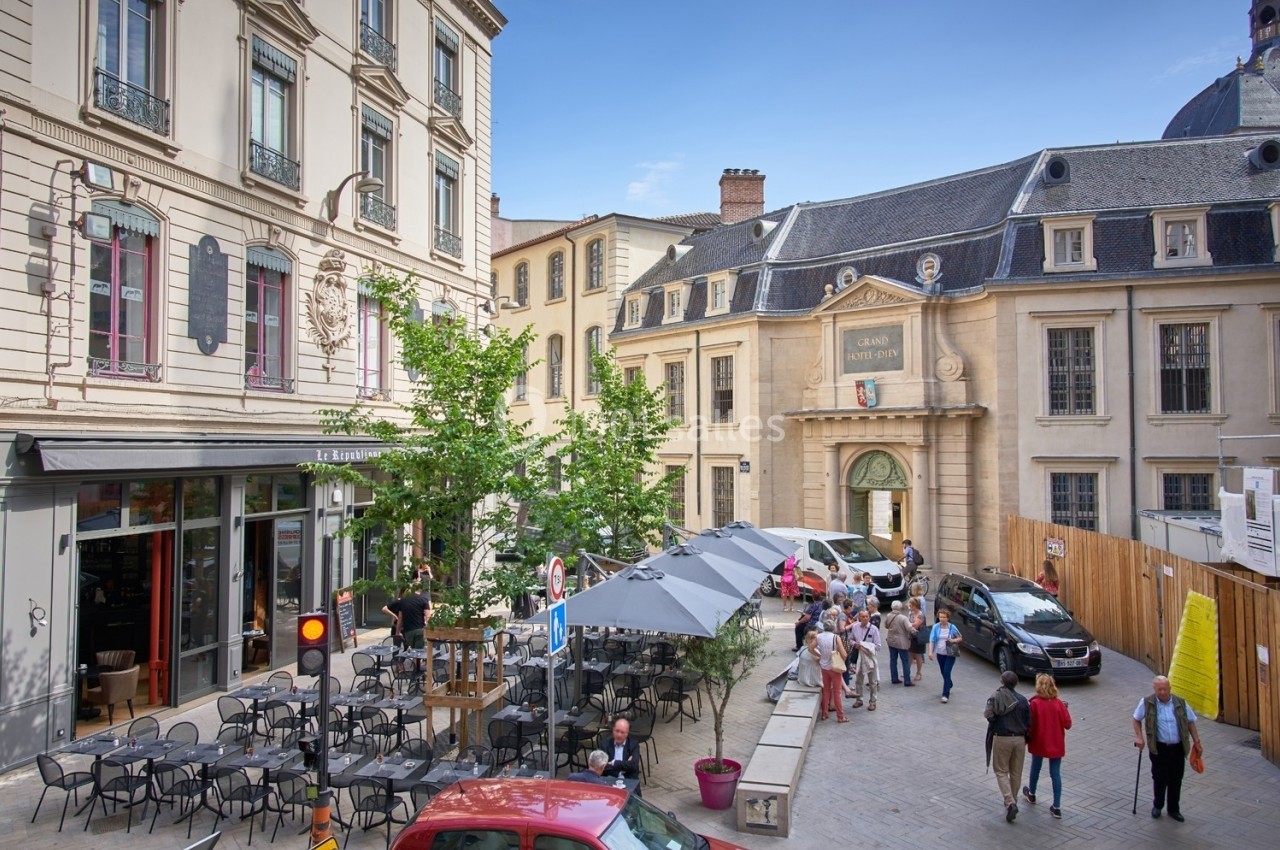 Place animée avec terrasse de café, piétons et bâtiments historiques sous un ciel dégagé.