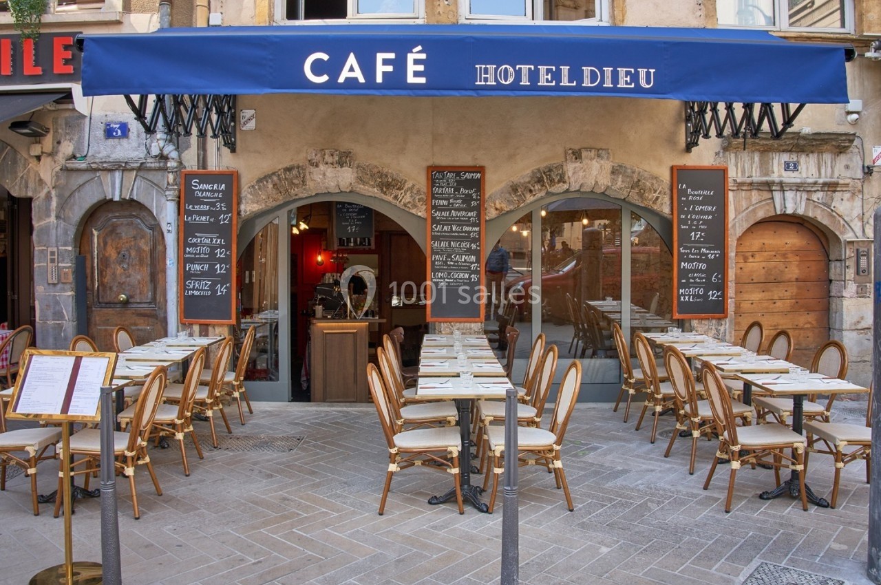 Terrasse d'un café avec tables et chaises alignées devant une façade en pierre, sous un auvent bleu.