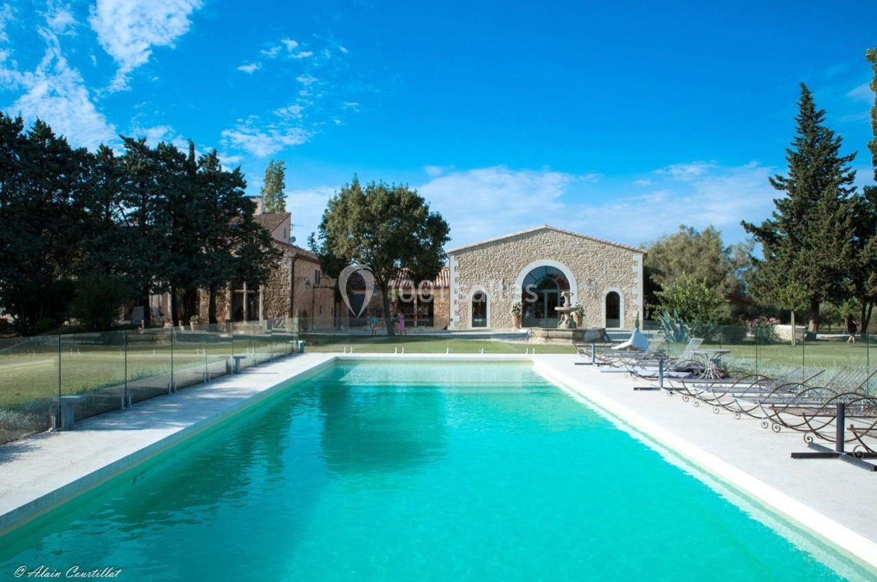 Piscine extérieure entourée de chaises longues, devant une maison en pierre avec jardin et arbres sous un ciel bleu.