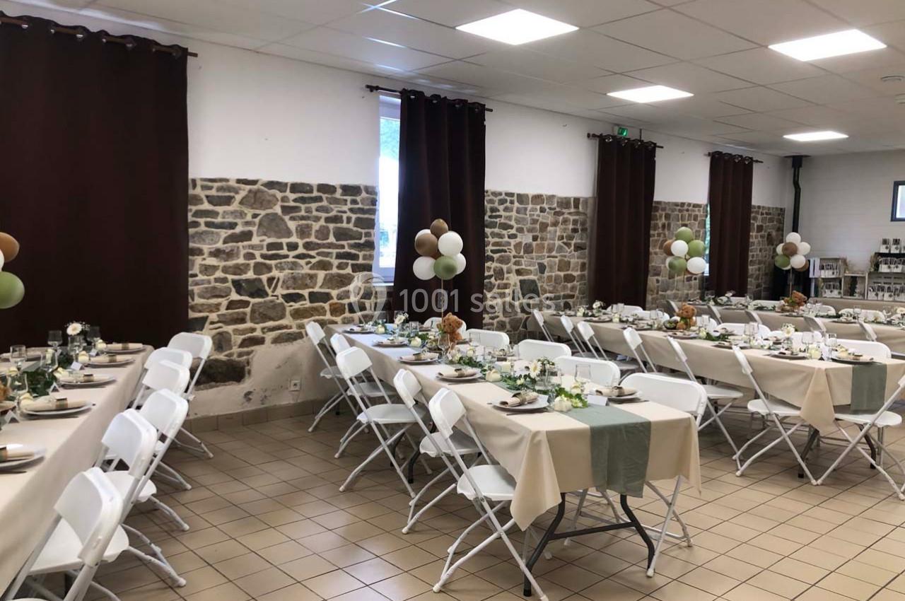 Salle décorée pour un événement avec des tables dressées, nappes beige et verte, chaises blanches et ballons en arrière-plan.