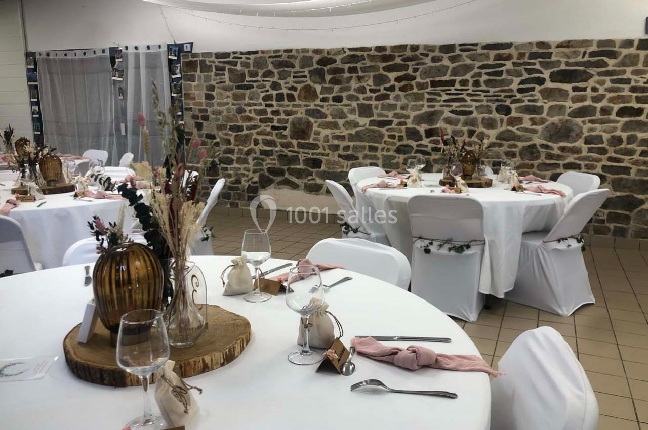 Salle décorée pour un événement avec des tables rondes dressées, nappes blanches et décorations florales rustiques.