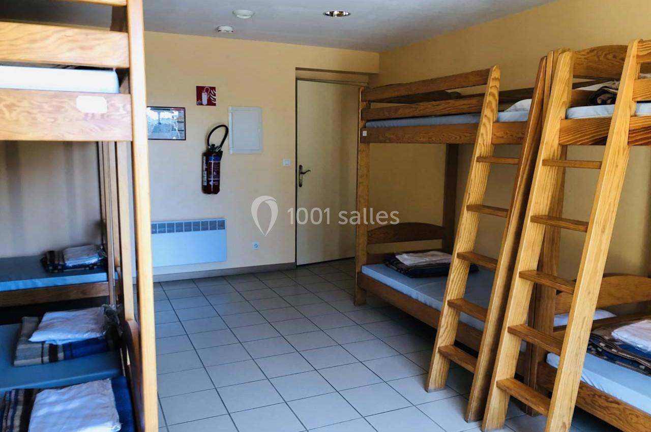 Dortoir avec lits superposés en bois, couvertures pliées, sol carrelé et mur beige, porte et extincteur visibles.