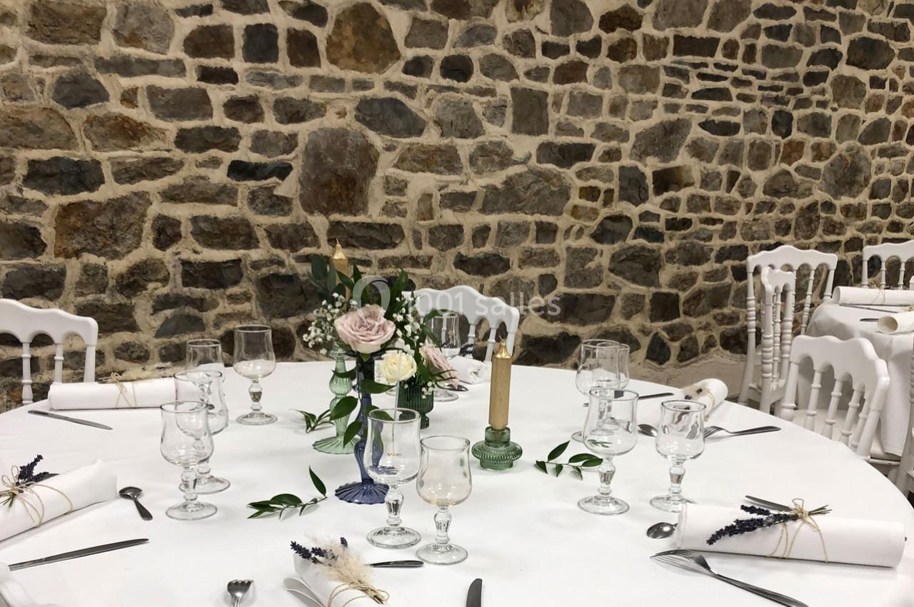 Table ronde décorée pour un événement, avec nappes blanches, verres, couverts, bougie et centre de table floral.
