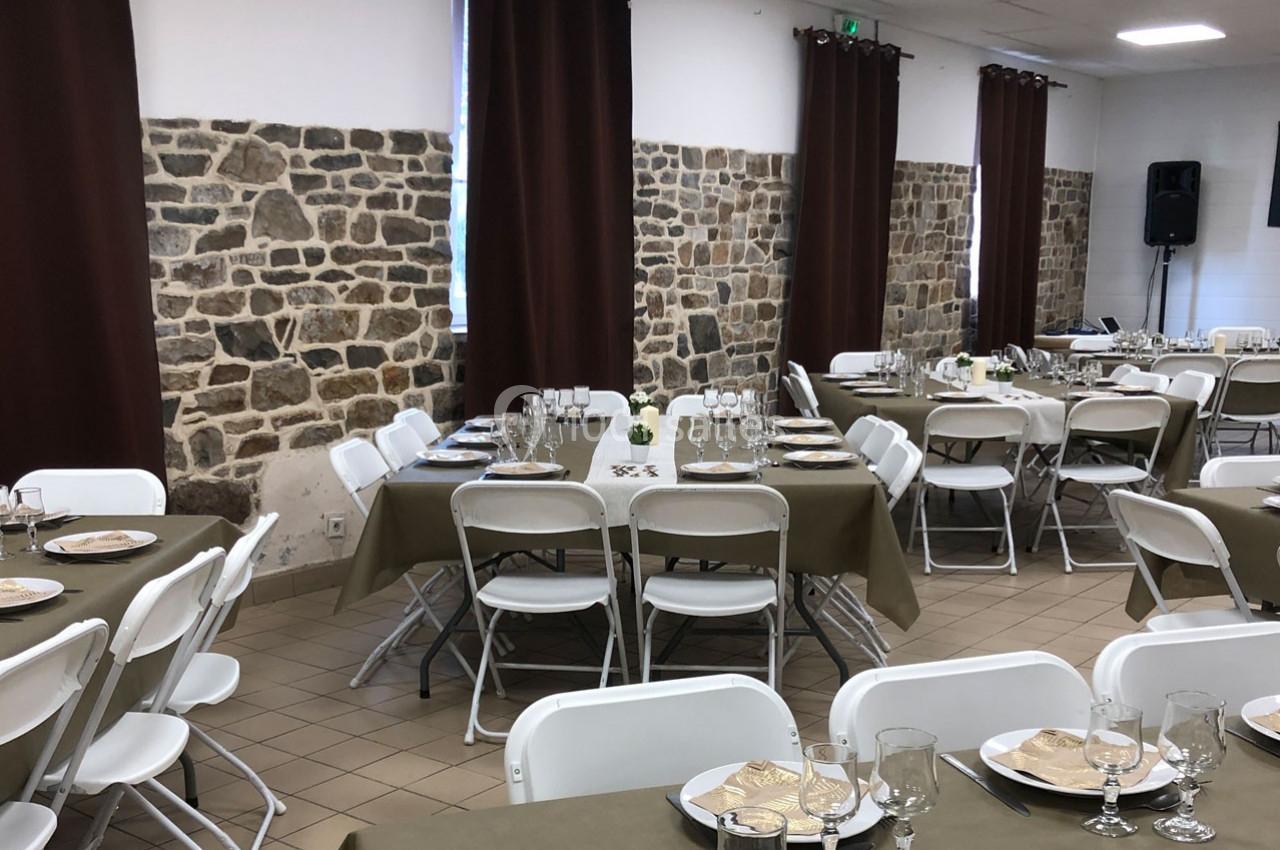 Salle décorée pour un repas, avec tables dressées, nappes brunes, chaises blanches et murs en pierre apparente.