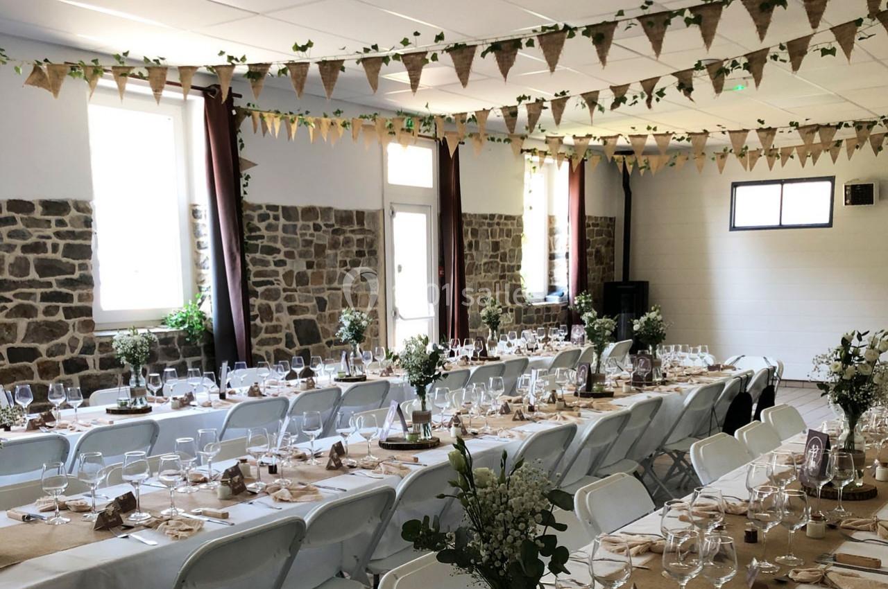Salle décorée pour un événement avec longues tables dressées, nappes en toile de jute, guirlandes et bouquets de fleurs.