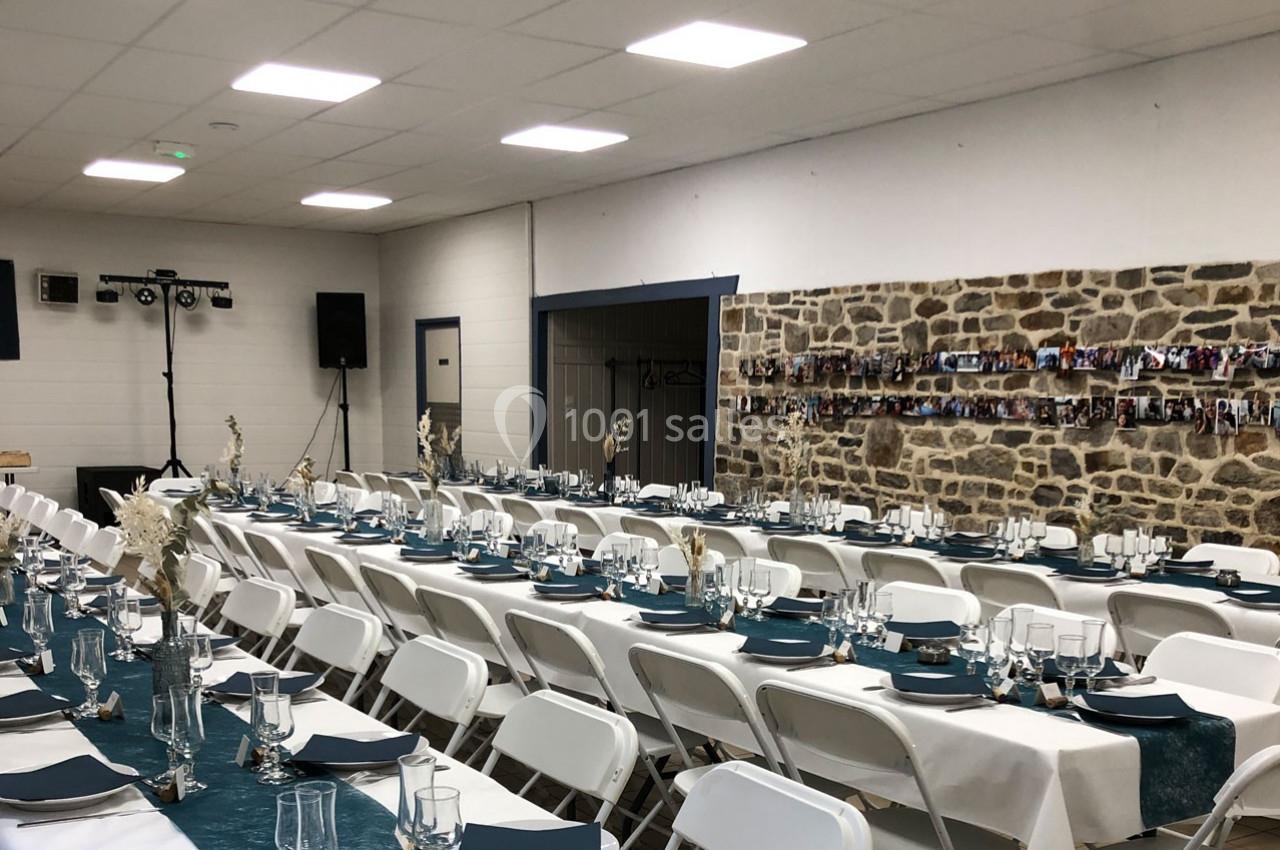 Salle décorée pour un événement avec des tables alignées, nappes blanches, chemins de table bleus et murs en pierre.