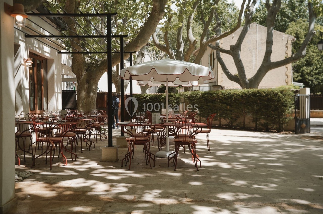 Terrasse d'un café avec des tables et chaises en métal rouge sous un parasol, entourée d'arbres et de lumière naturelle.