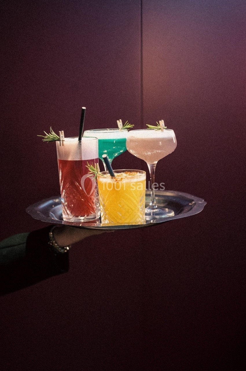Plateau avec quatre cocktails colorés garnis de brins de romarin, tenu par une main sur un fond sombre.