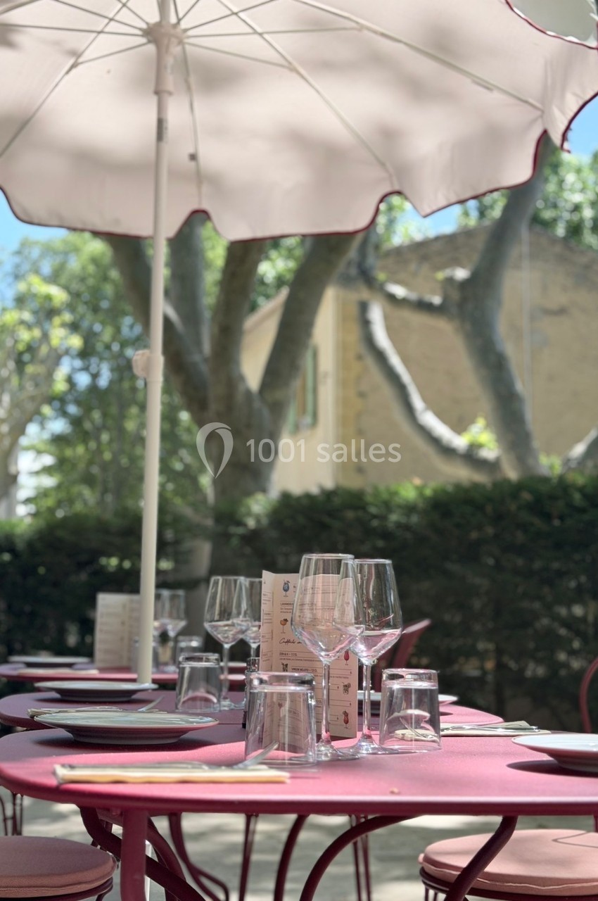 Table de restaurant en extérieur avec parasol, verres et couverts, entourée de verdure et d'arbres sous un ciel clair.