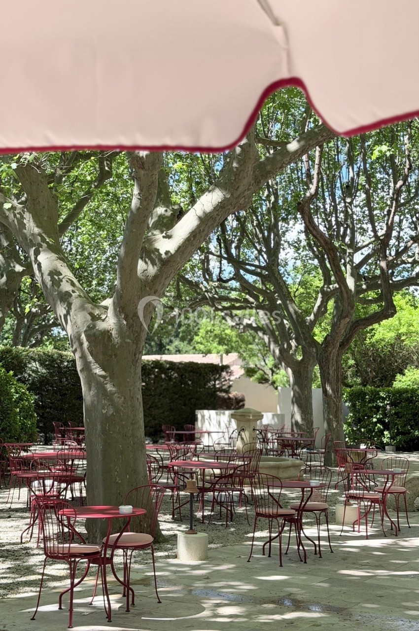 Terrasse ombragée avec des tables et chaises rouges sous de grands arbres, partiellement couverte par un parasol beige.