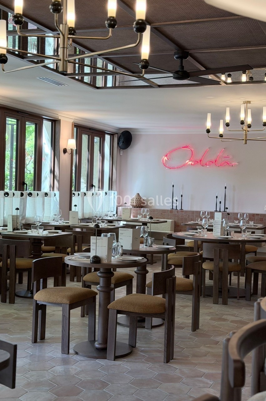 Salle de restaurant lumineuse avec tables en bois, chaises rembourrées et enseigne néon rose au mur.